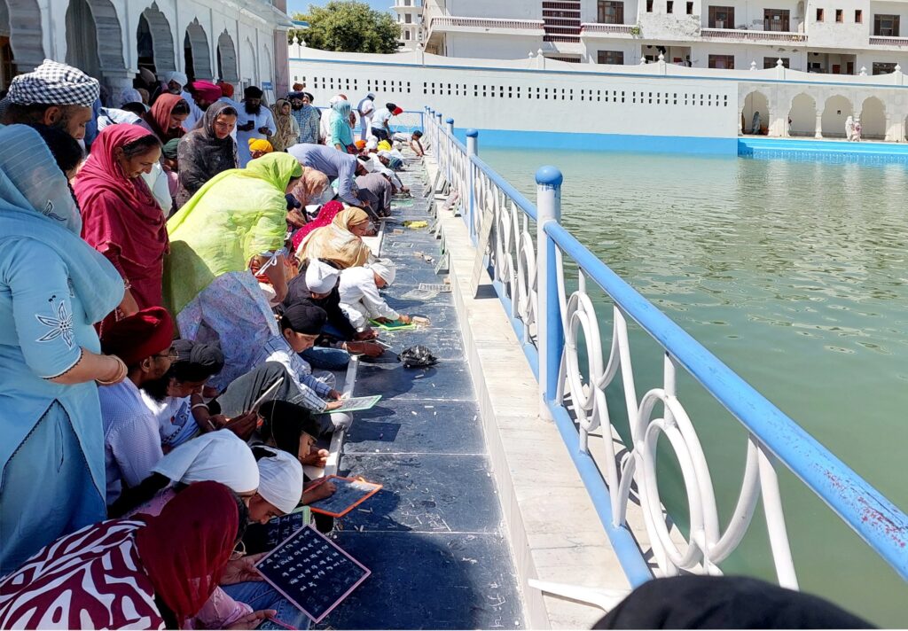 Children write Gurmukhi alphabets on slates in Gurdwara Likhansar Sahib, facing the Sarovar.